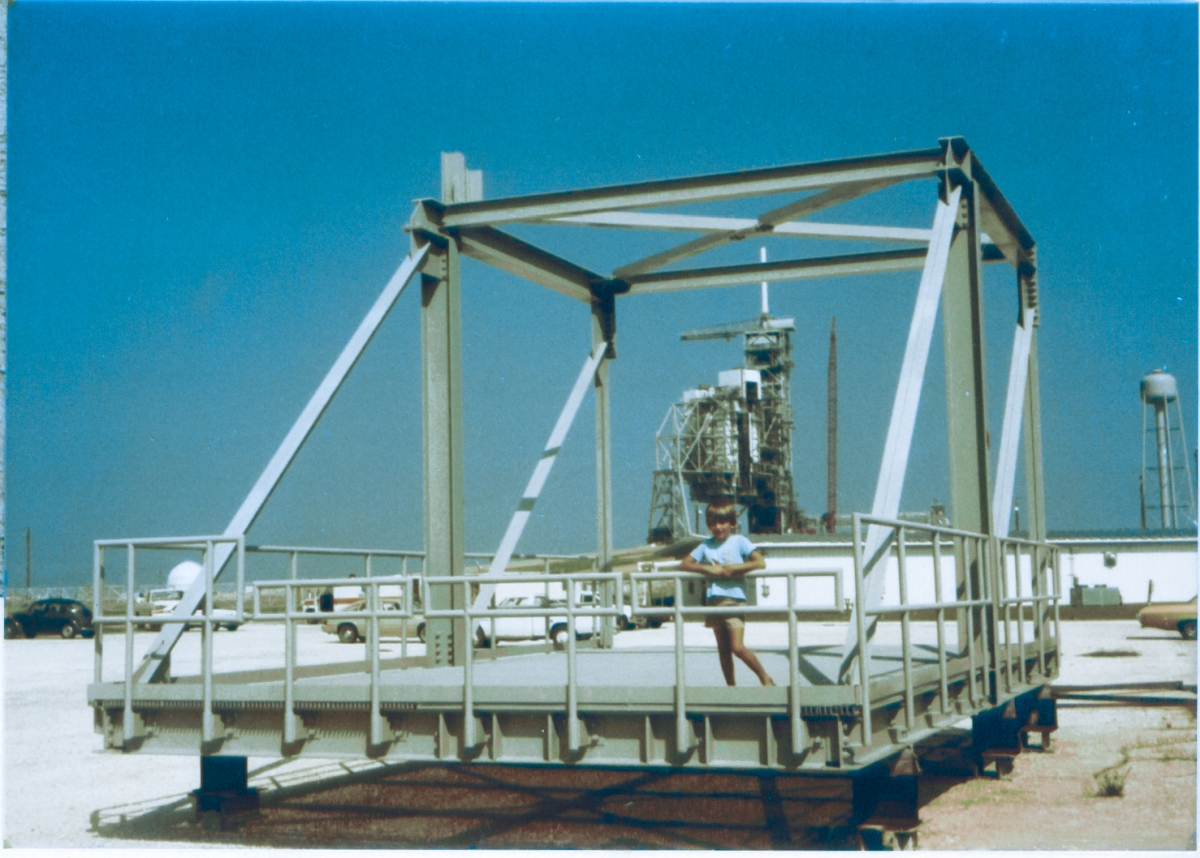 A young Kai MacLaren stands on the Centaur Porch with the pad structures at Space Shuttle Launch Complex 39-B over his head, in the distance directly behind him. The Centaur Porch was designed and built to support part of the Centaur Rolling Beam Umbilical Assembly, and was later lifted into place and bolted on to the structure in the distance behind Kai. Kai is standing on actual launch pad hardware, and he knows it. We had a very good day together at Pad B, this day.